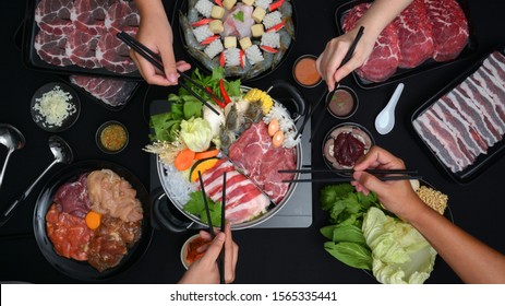 Top View Of People Eating Shabu-Shabu In Hot Pot With Fresh Sliced Meat, Sea Food, And Vegetables With Black Background, Japanese Cuisine