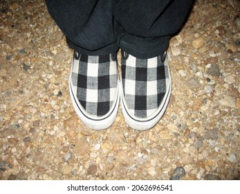 Top View Of A Person Wearing Black Jeans With A Pair Of Checkerboard Style Slip-on Shoes Standing On Pebble Floor