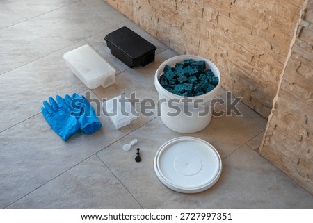 Top view of pest control kit including blue bait blocks, plastic trap, and gloves on a tiled floor.
