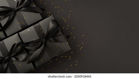 Top view photo of black gift boxes with black ribbon bow tag and golden confetti on isolated black background with blank space