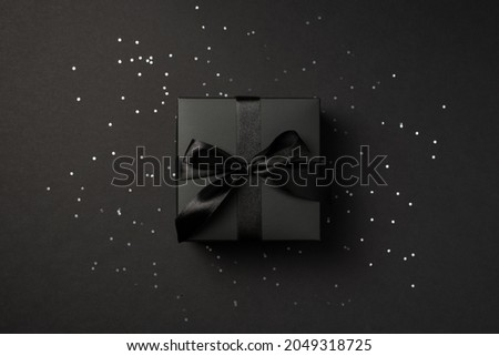 Top view photo of black giftbox with black satin ribbon bow over shiny sequins on isolated black background