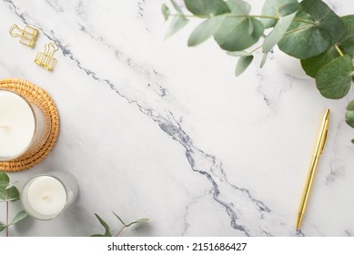 Top view photo of candles on rattan serving mat gold pen binder clips and vase with eucalyptus on white marble background with copyspace