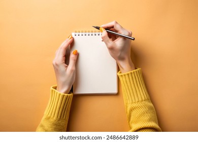 Top view photo of female hands in yellow pullover writing in spiral notepad on isolated light orange background with copyspace