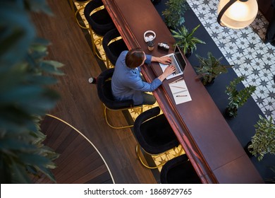 Top View Photo Of A Focused Gentleman Sitting On Stool While Typing On Laptop At A Fancy Hotel Lounge