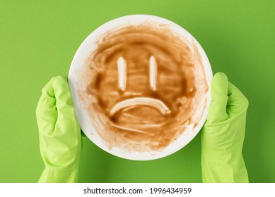 Top View Photo Of Hands In Green Rubber Gloves Holding Dirty Dish With Sad Face On Isolated Green Background