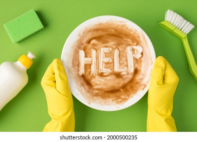 Top View Photo Of Hands In Yellow Rubber Gloves Holding Dirty Dish With Inscription Help White Detergent Gel Bottle Green Brush And Scouring Pad On Isolated Green Background