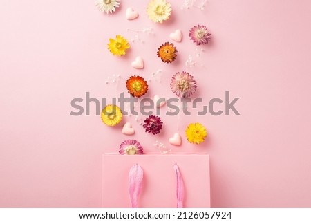 Top view photo of the pink shopping bag many colorful flowers with branches of gypsophila and pink confetti in shape of hearts scattered above bag on the pastel pink isolated background blank space