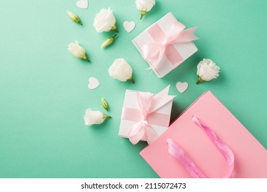 Top view photo of two small white gift boxes with cute silk pink bows white eustomas and small confetti in shape of hearts near the big pink shopping bag on the pastel turquoise background