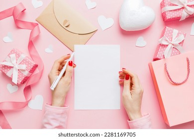 top view photo of valentine day decor female hands holding letter with envelope, small gift box and heart decoration on isolated pastel colored background with empty space.