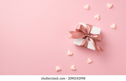 Top view photo of valentine's day decorations white giftbox with pink silk ribbon bow and small hearts on isolated pastel pink background with copyspace
