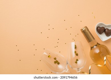 Top view photo of valentine's day decor heart shaped saucer with chocolate candies wine bottle two wineglasses with confetti and sequins on isolated pastel beige background with copyspace