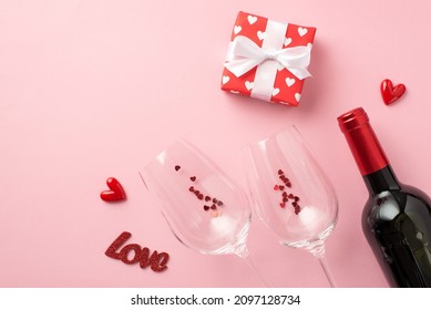 Top view photo of valentine's day decor hearts giftbox in wrapping paper with pattern of hearts inscription love wineglasses with confetti wine bottle on isolated pastel pink background with copyspace