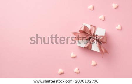 Top view photo of valentine's day decorations white giftbox with pink silk ribbon bow and small hearts on isolated pastel pink background with copyspace
