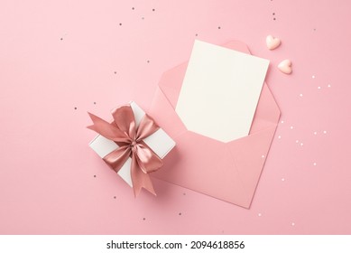 Top view photo of white giftbox with pink ribbon bow small hearts open pink envelope with paper card and shiny sequins on isolated pastel pink background with blank space
