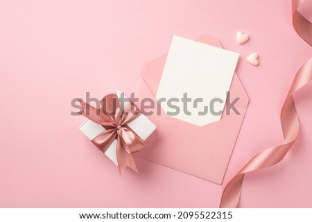 Top view photo of white giftbox with pink bow small hearts curly silk ribbon and open pink envelope with paper card on isolated pastel pink background with blank space