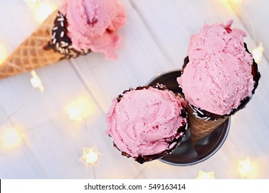 Top View Of Pink Strawberry Flavor Ice Cream In Waffle Cones Shot Over A Rustic Background With Fairy Lights. Extreme Shallow Depth Of Field With Selective Focus On Two Desserts In The Foreground.