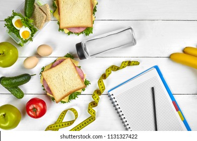Top view planning notebook with copy space and healthy breakfast. Sandwich with ham and cheese, eggs, cucumber, tomato and bottle of water on white wooden table.