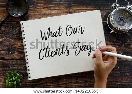 Top view of plant,magnifying glass,clock and hand holding pen writing ' What Our Clients Say ' on notebook on wooden background.