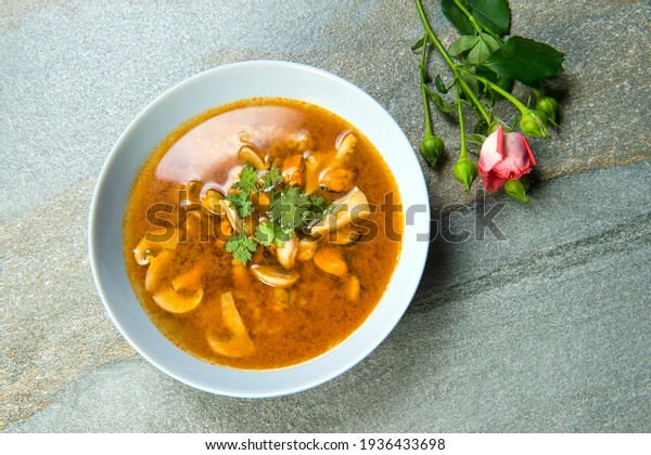 Top view of a plate with Pan Asian traditional\
seafood soup with mussels, calamari, mushrooms served with fresh\
parsley herbs on gray stone background with pink rose flower brunch\
near dish