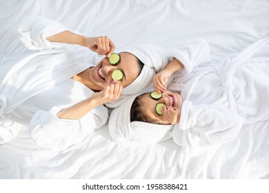 Top view of playful mother and daughter in bathrobes and white towels on heads laying on bed, holding cucumber circles, making face care procedures at home. Motherhood, parenthood concept - Powered by Shutterstock