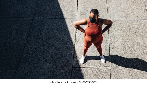 Top View Of A Plus Size Woman In Sportswear And Protective Face Mask Standing With Hands On Hips. Female Taking Break From Workout Outdoors.