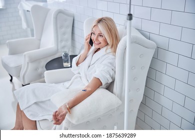 Top view portrait of blonde Caucasian pretty female in white plush bathrobe sitting in style chair while receiving treatment vitamin drip in medicine clinic