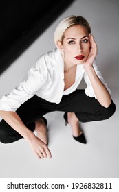 Top View Portrait Of Daring Trendy Woman Posing On White Studio Background Sitting On Floor. Gorgeous Stylish Blonde Female Wearing White Shirt, Black Pants And High Heel Shoes Looking To Camera