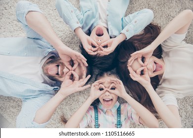 Top View Portrait Of Foolish Comic Family With Two Kids Lying On Carpet Making Eyewear With Fingers Gesturing Tongue-out Happy Together. Freetime Daydream Concept