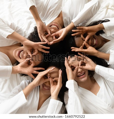 Top view portrait funny diverse girls wearing white bathrobes lying in bed, making binoculars with hands, looking at camera, five beautiful young women posing, celebrating hen party, square photo