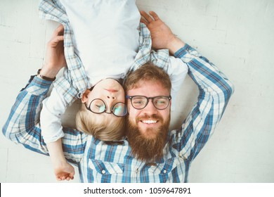 Top View Portrait Of Happy Beard Father And Son In Eyeglasses Lying On The White Floor Background. Happy Family Spend Time Together. Father's Day Concept.