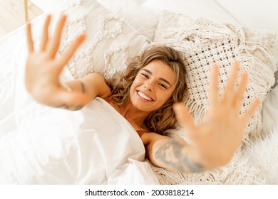 Top-View-Porträt von glücklicher fröhlicher Frau liegt in weißem, gemütlichem Bett aufwachen am Morgen, zieht die Hände nach vorn. Warme Farben, boho-Interieur. 