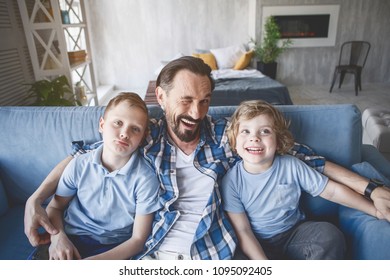 Top View Portrait Of Happy Dad Winking While Hugging Kids On Couch In Apartment