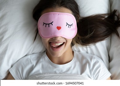 Top view portrait of happy girl lying in comfortable bed wake up in morning have fun making funny faces, smiling young woman in eye sleeping mask awaken in cozy bedroom feel optimistic at home
