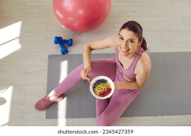 Top view portrait of a happy smiling sporty slim woman wearing sportswear eating healthy food after working sitting on yoga mat. Fit girl with a bowl of fruits and berries. Healthy nutrition concept. - Powered by Shutterstock