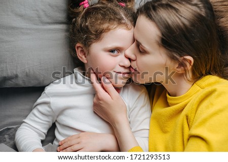 Top view portrait of happy young mother kissing cute little kid girl hugging bonding lying on bed, young cheerful loving mom and daughter laugh spend time together at home