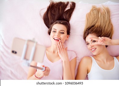 Top View Portrait Of  Joyful, Attractive Girls With Blonde, Brunette Hair Laying On Bed On Back Shooting Selfie On Front Camera Using Smart Phone, Selfish Stick, Gesture Peace Symbol Near Eye