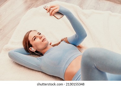 Top view portrait of relaxed woman listening to music with headphones lying on carpet at home. She is dressed in a blue tracksuit. - Shutterstock ID 2130155348