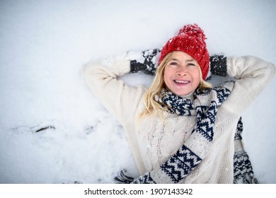 Top View Portrait Of Senior Woman With Hat Outdoors Lying On Snow, Looking At Camera.