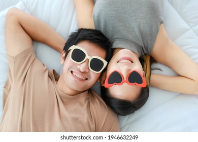Top view portrait shot of romantic young Asian lover couple cute smiling wearing stylish modern sunglasses, lying on the bed and looking at the camera. Concept of couple honeymoon in summer