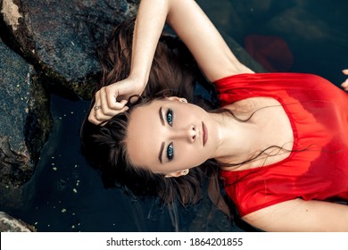 Top view portrait of young pretty wet woman mermaid lying on rock in the water river or lake touching head, dressed in red dress, looking at the camera, copy space and nature blur background.
