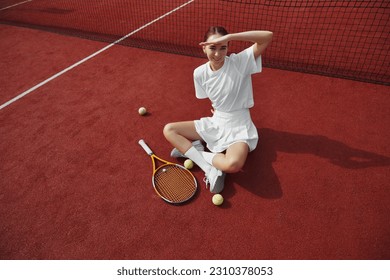 Top view portrait of young woman in white outfit covering face by hand from sunlight while sitting on floor in outdoor tennis court, taking break from practice. - Powered by Shutterstock