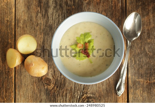 Top view of a potato soup in a\
blue bowl with fresh potatoes and spoon on a wooden\
background