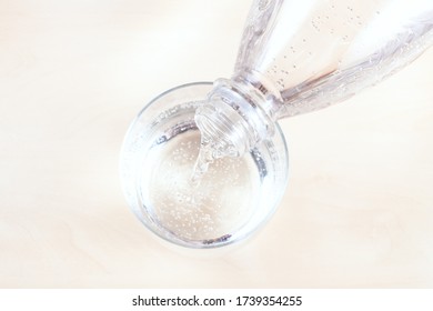 Top View Of Pouring Sparkling Mineral Water From Transparent Plastic Bottle In Glass On Light Brown Table Close Up