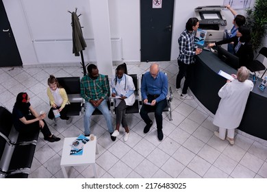 Top View Of Private Clinic Front Desk With Asian Patient Registering For Doctor Appointment. Diverse Group Of Patients Wait Sitting In Hospital Lobby For Clinical Consult In Hospital Waiting Area.