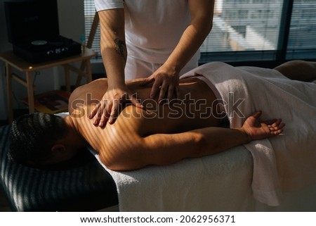Top view of professional male masseur with strong tattooed hands massaging back and shoulders of muscular sports man lying on stomach, at massage table in luxury salon, on background of sunlight.