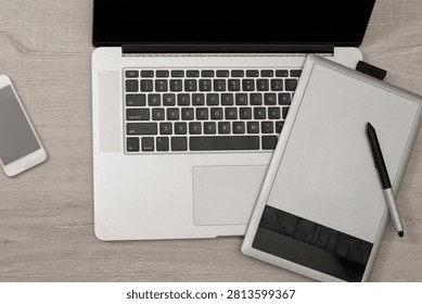 Top view of a professional workspace featuring a modern laptop, a digital drawing tablet with a stylus, and a smartphone placed on a light wooden desk surface.