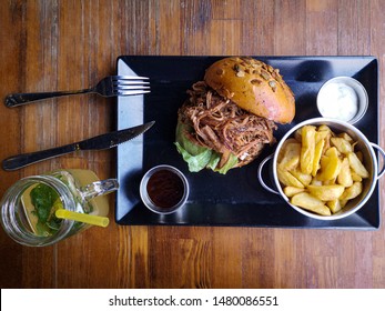 Top View Of Pulled Pork Sandwich With Fried Wedges Red And White Sauce, Fork, Knife And A Lemonade
