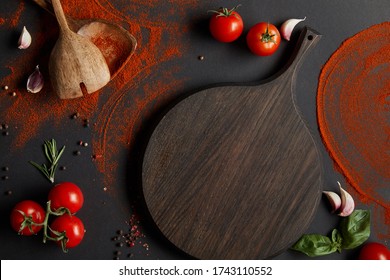 top view of red cherry tomatoes, cutting board, garlic cloves and fresh herbs near spoons and paprika powder on black