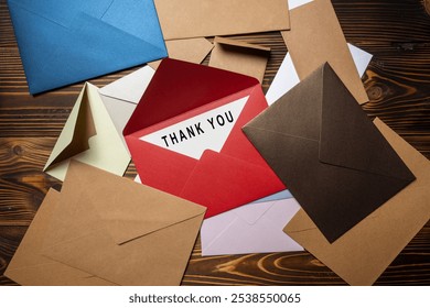 Top view of red envelope and postcard with white card with words Thank You written on it on stack of envelope and postcard on wooden background. Thank you message for presentation or business