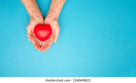 Top View Of A Red Heart On The Palm Senior Woman Over A Light Blue Background. World Heart Day, World Health Day. Organ Donation. People, Age, Love, Praying, And Health Care Concept